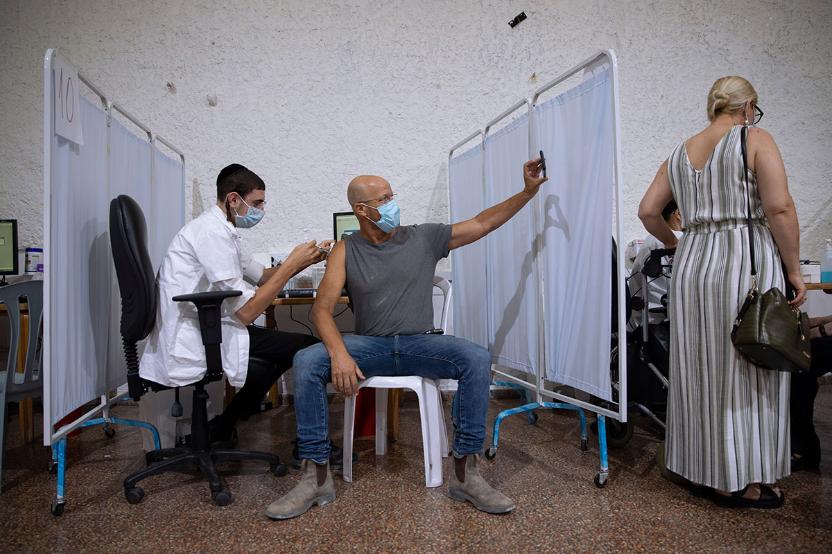 [Symbolbild] Ein Israeli (Mitte) macht am 30. August 2021 ein Selfie, während er von medizinischem Personal in einem Coronavirus-Impfzentrum in Ramat Gan, Israel, eine dritte Impfdosis gegen COVID-19 erhält. Rückschlüsse auf Impfnebenwirkungen können nicht aus der Statistik über die Verstorbene an einer Coivd-19-Infektion gezogen werden. Foto: KEYSTONE/ AP Photo/ Oded Balilty